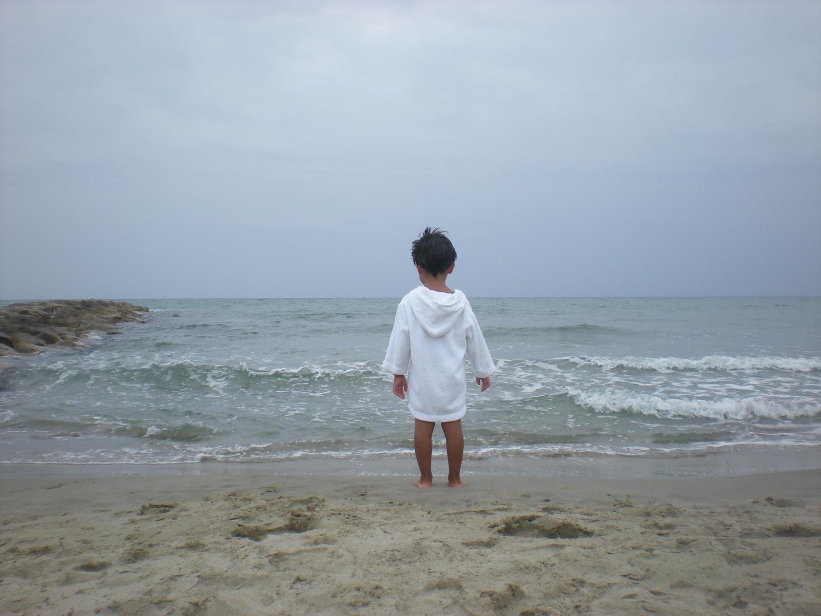 Un enfant vêtu de blanc est debout face à la mer par temps gris
