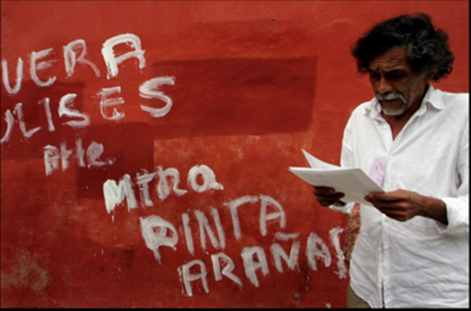 Devant un mur rouge aux inscriptions blanches, un homme barbu lit des feuillets en papier