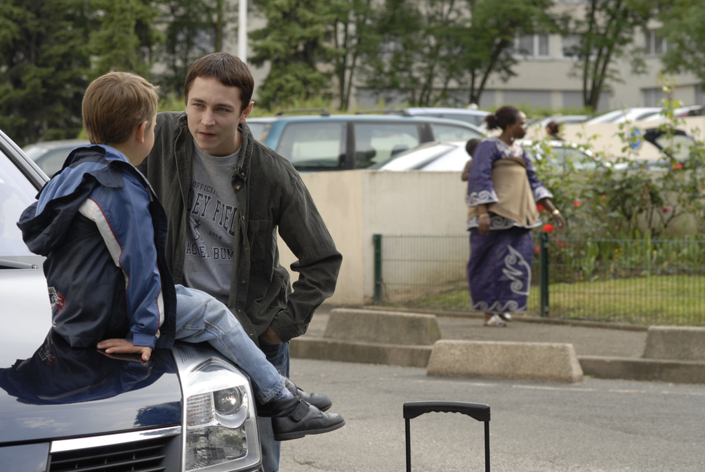 //www.occitanie-films.netPetit garçon assis sur le capot avant d'une voiture, parlant avec un adolescent