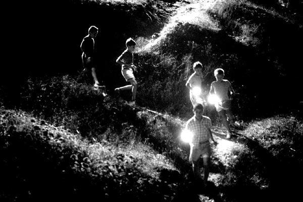 Photo de nuit, en noir et blanc, d'un groupe d'enfants se promenant dans la nature, éclairant leur chemin à la lampe torche